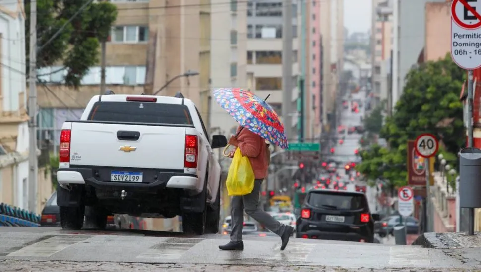 imagem mostra uma caminhonete branca e uma pessoa com um guarda-chuva colorido atravessando a rua.