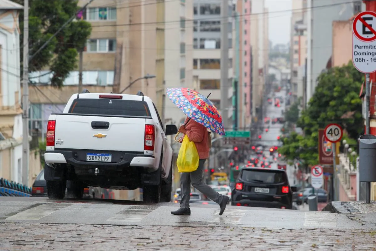 imagem mostra uma caminhonete branca e uma pessoa com um guarda-chuva colorido atravessando a rua.