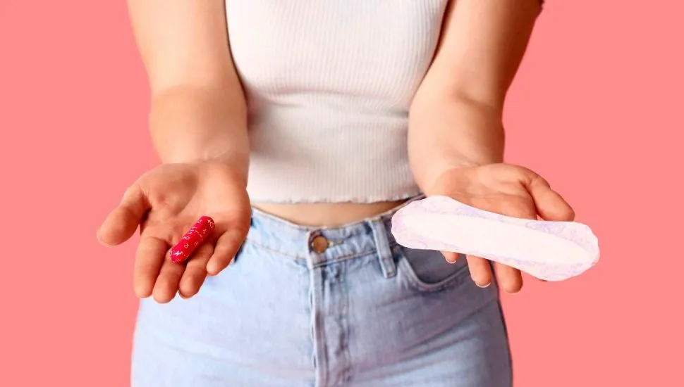 Imagem mostra uma mulher de calça jeans e camiseta branca segurando um absorvente.