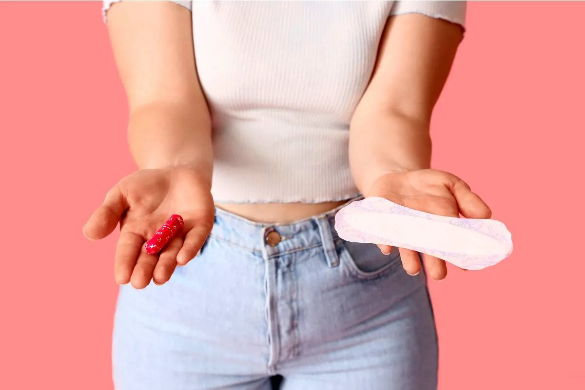Imagem mostra uma mulher de calça jeans e camiseta branca segurando um absorvente.