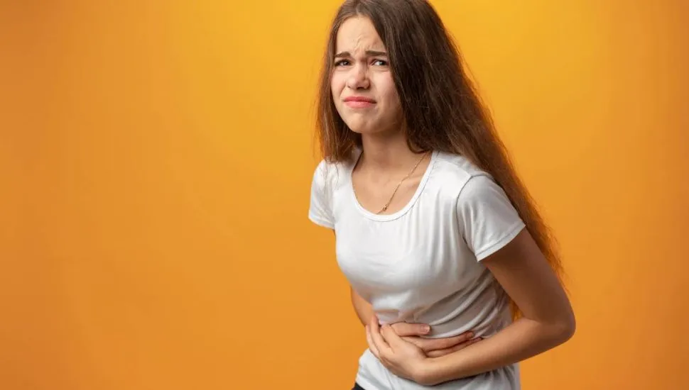 Imagem mostra uma mulher com dor de barriga. Ela esta em um fundo amarelo, com camiseta branca e calça preta.