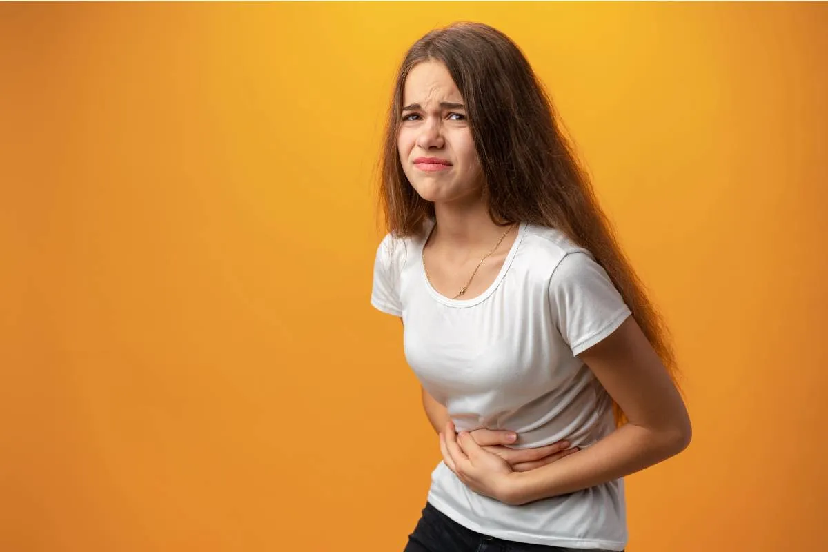 Imagem mostra uma mulher com dor de barriga. Ela esta em um fundo amarelo, com camiseta branca e calça preta.