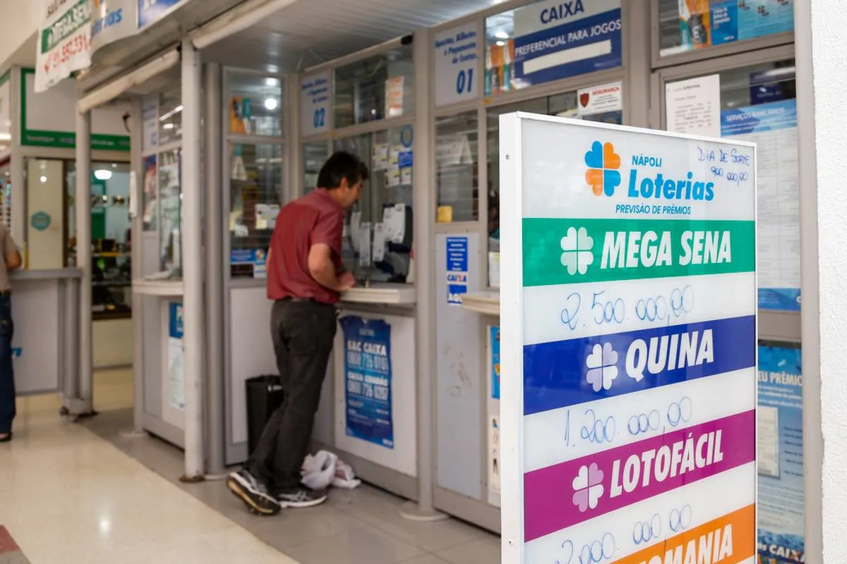 Imagem mostra uma pessoa apostando na loteria dentro de uma lotérica. Na imagem tem um homem de calça preta e camiseta vermelha no guichê de atendimento.