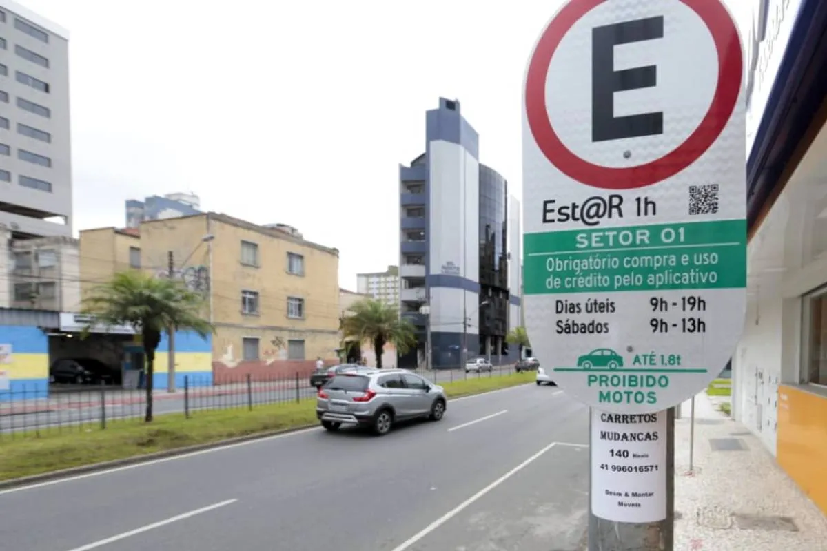 Imagem mostra uma placa de transito na beira de uma rua em Curitiba. O céu está fechado e tem um carro andando na rua.