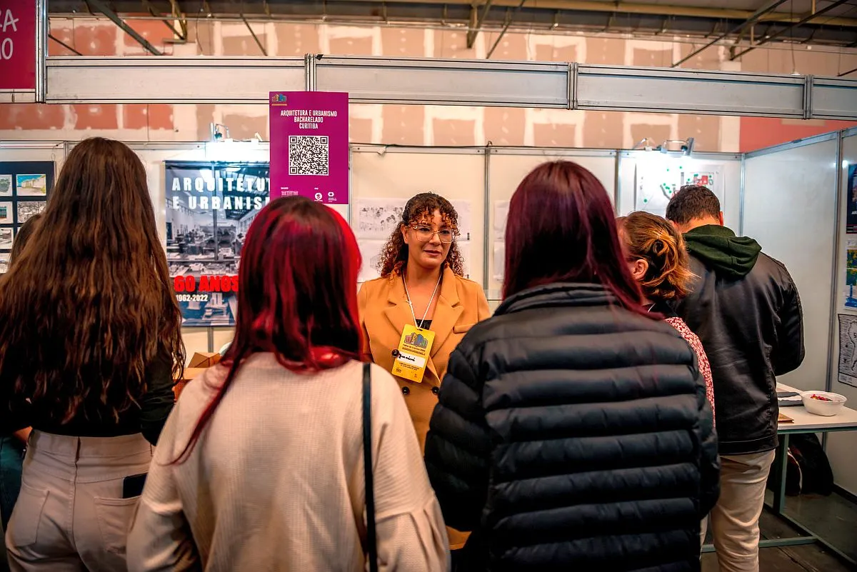 Foto mostra expositora diante de um stand falando para quatro estudantes durante edição anterior da feira