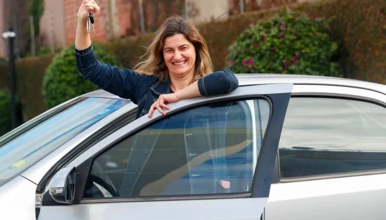 Imagem mostra uma mulher na porta de um carro cinza. Ela sorri e mostra a chave de um carro.
