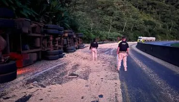 Imagem mostra um caminhão tombado e muita carga na pista.