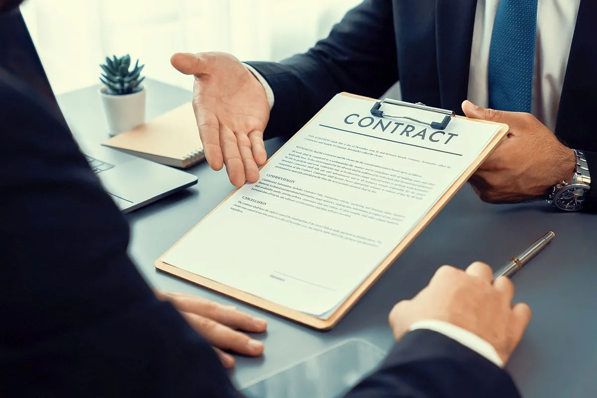 Imagem mostra dois homens sentados à mesa com um contrato nas mãos negociando