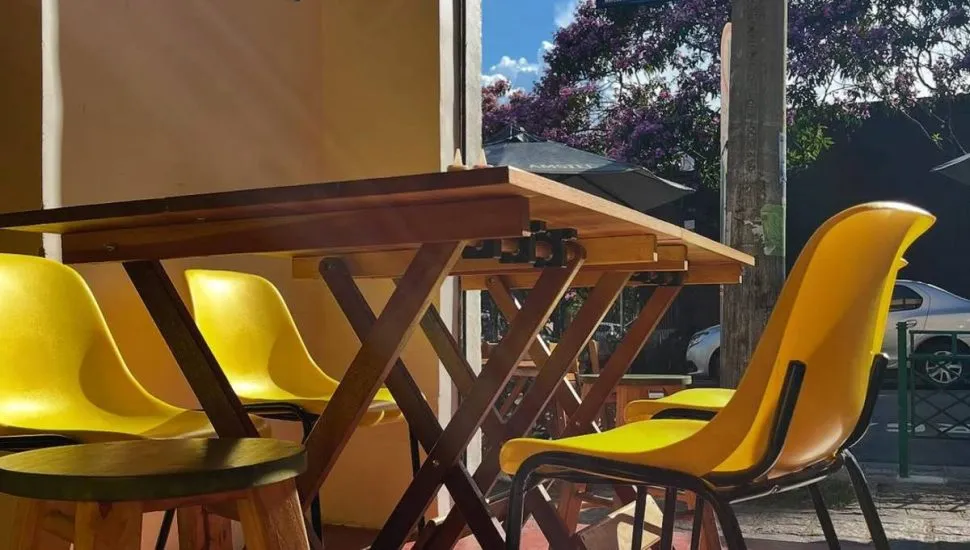 Imagem mostra cadeiras de um bar de Curitiba. Elas são de madeira com detalhes em amarelo. O sol bate e ilumina a mesa. Ao fundo, árvores e uma placa azul de nome de rua.