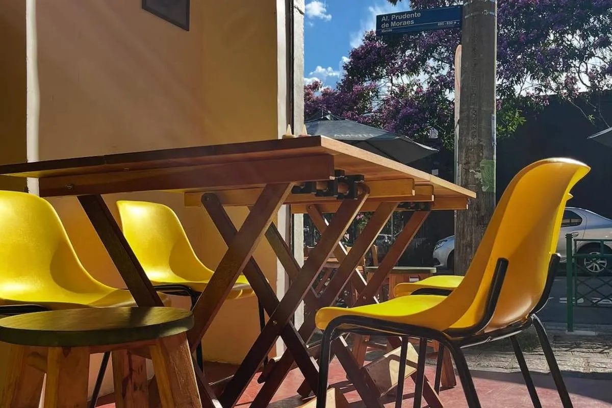 Imagem mostra cadeiras de um bar de Curitiba. Elas são de madeira com detalhes em amarelo. O sol bate e ilumina a mesa. Ao fundo, árvores e uma placa azul de nome de rua.