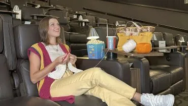 Imagem mostra uma mulhar sentada na poltrona d eum cinema, fazendo crochê, com um balde de pipoca e uma bolsa com apetrechos de crochê. a sala de está vazia