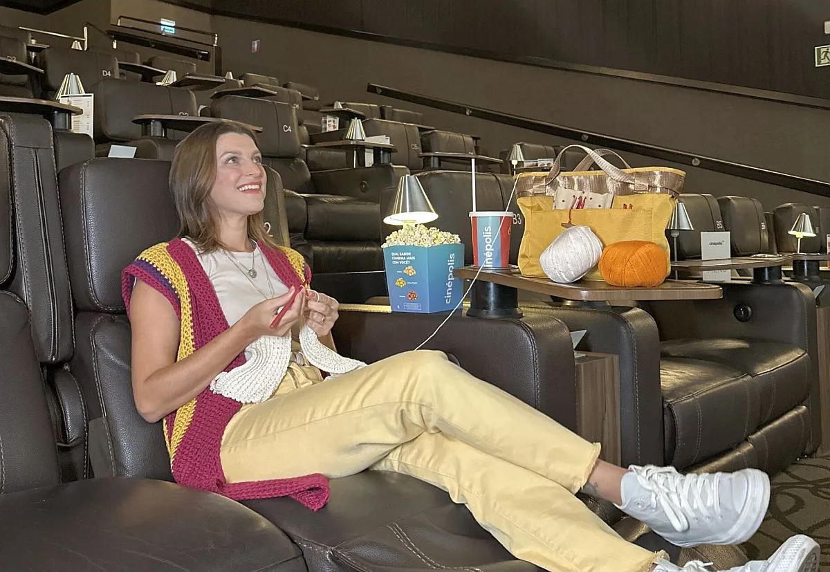 Imagem mostra uma mulhar sentada na poltrona d eum cinema, fazendo crochê, com um balde de pipoca e uma bolsa com apetrechos de crochê. a sala de está vazia