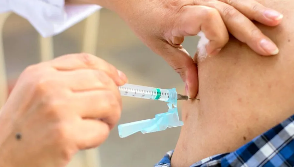 Imagem mostra em close as mãos de um enfermeiro vacinando uma pessoa que veste uma camisa xadrez no braço