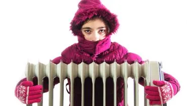 Imagem mostra uma mulher com uma blusa pesada, luva e gorro abraçando um aquecedor.