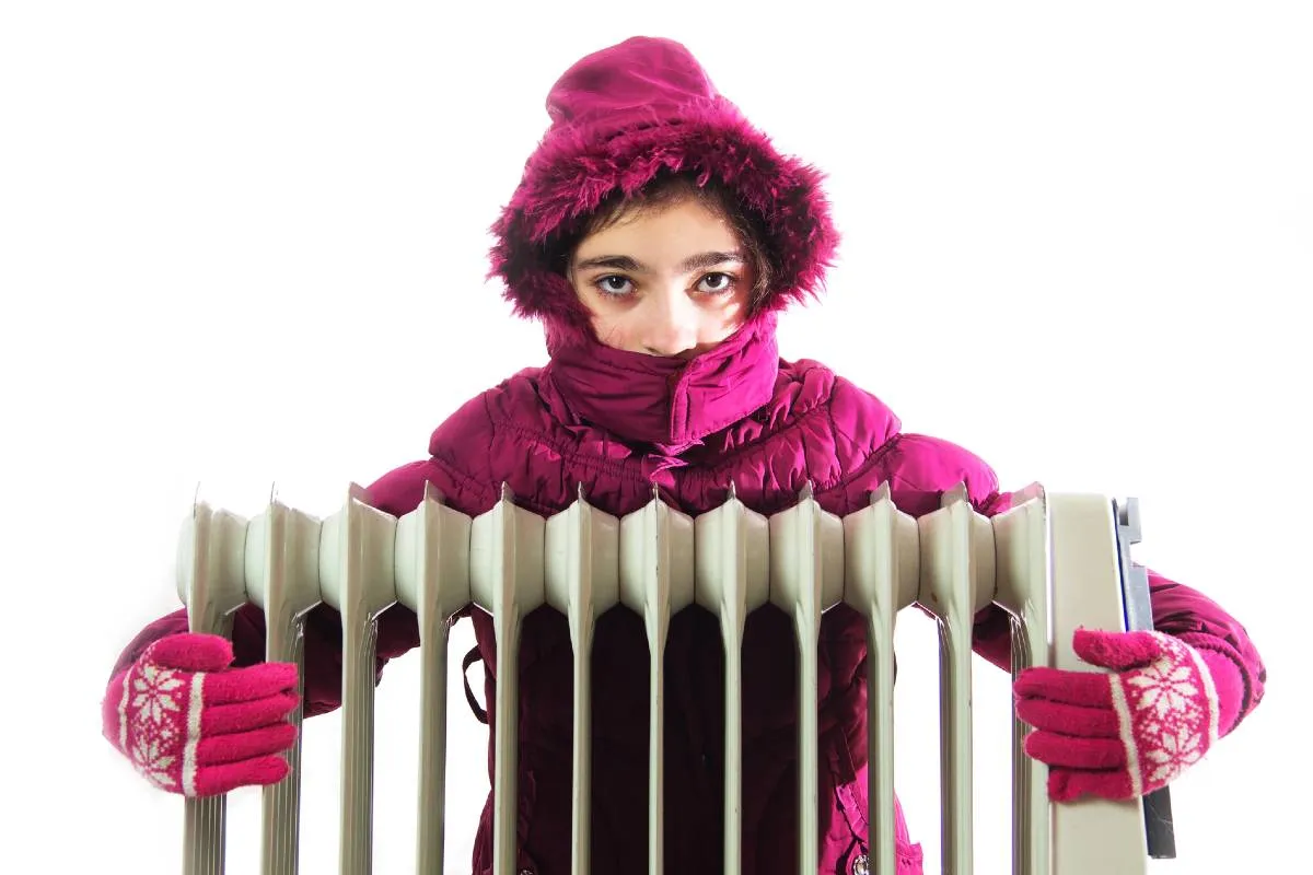 Imagem mostra uma mulher com uma blusa pesada, luva e gorro abraçando um aquecedor.