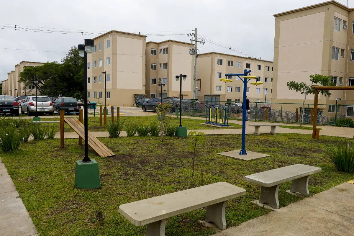 Imagem mostra um prédio da Cohab em Curitiba com céu nublado e um parquinho.