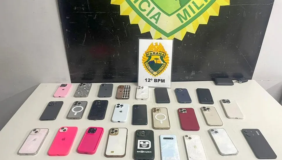 Dezenas de celulares de diversas marcas estão sobre uma mesa em frente ao logotipo da Polícia Militar do Paraná.