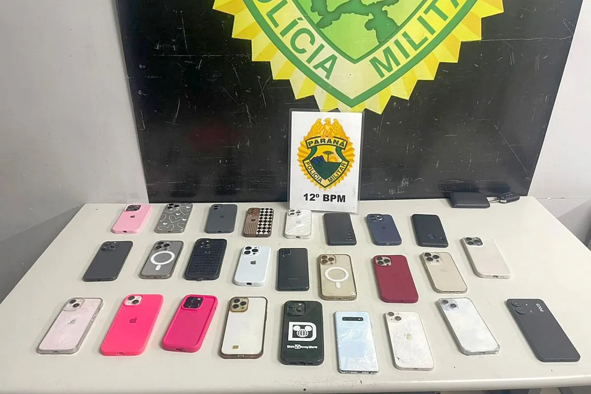 Dezenas de celulares de diversas marcas estão sobre uma mesa em frente ao logotipo da Polícia Militar do Paraná.