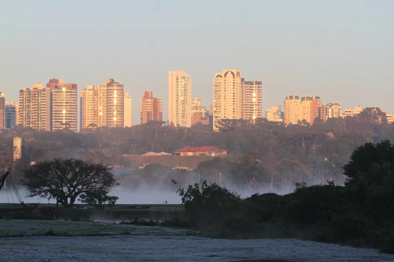 Imagem mostra prédios de Curitiba ao fundo, com lago do Parque Barigui em segundo plano e gramado com geada em primeiro plano