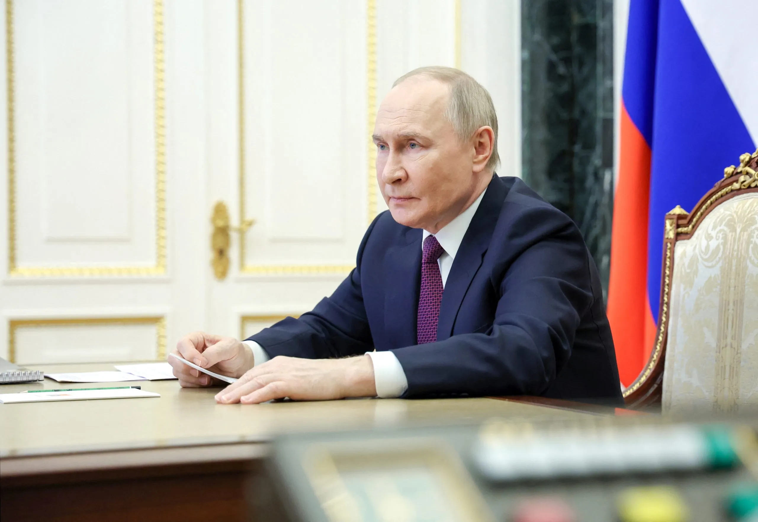 Presidente russo vladmir putin sentado à mesa, com uam folha de papel nas maos a ao lado de uma bandeira russa