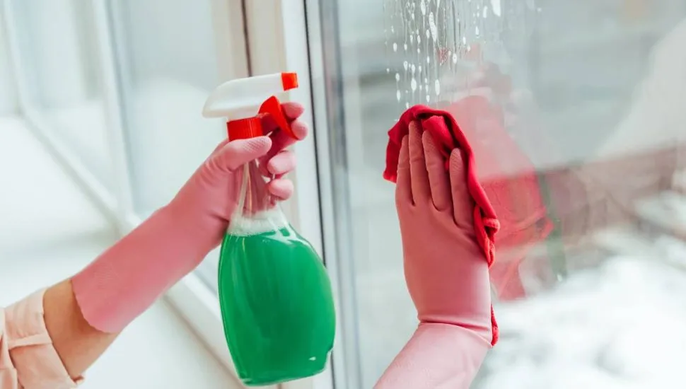 Imagem mostra uma pessoa limpando um vidro com um pano vermelho e um borrifador com um líquido verde.