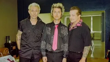 Imagem mostra os integrantes do Green Day. Eles estão de preto e um deles, o vocalista, usa uma gravada rosa.