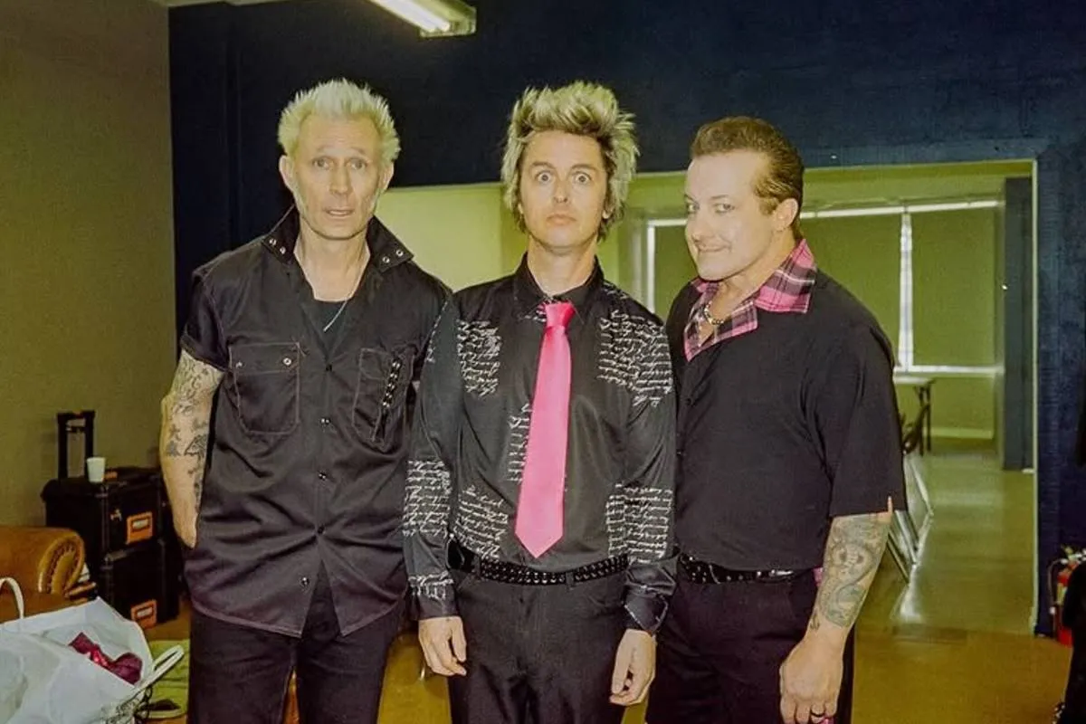 Imagem mostra os integrantes do Green Day. Eles estão de preto e um deles, o vocalista, usa uma gravada rosa.