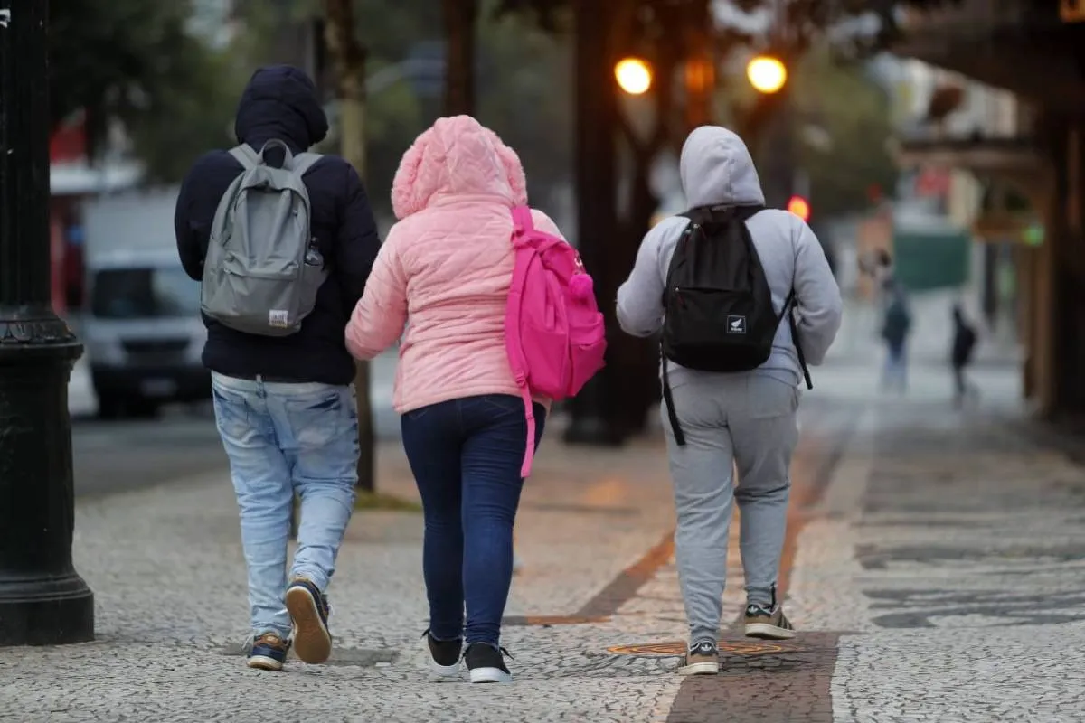 Imagem mostra três pessoas com muito frio em Curitiba. Eles estão encolhidos, com os braços entrelaçados, capuz e muuuito frio