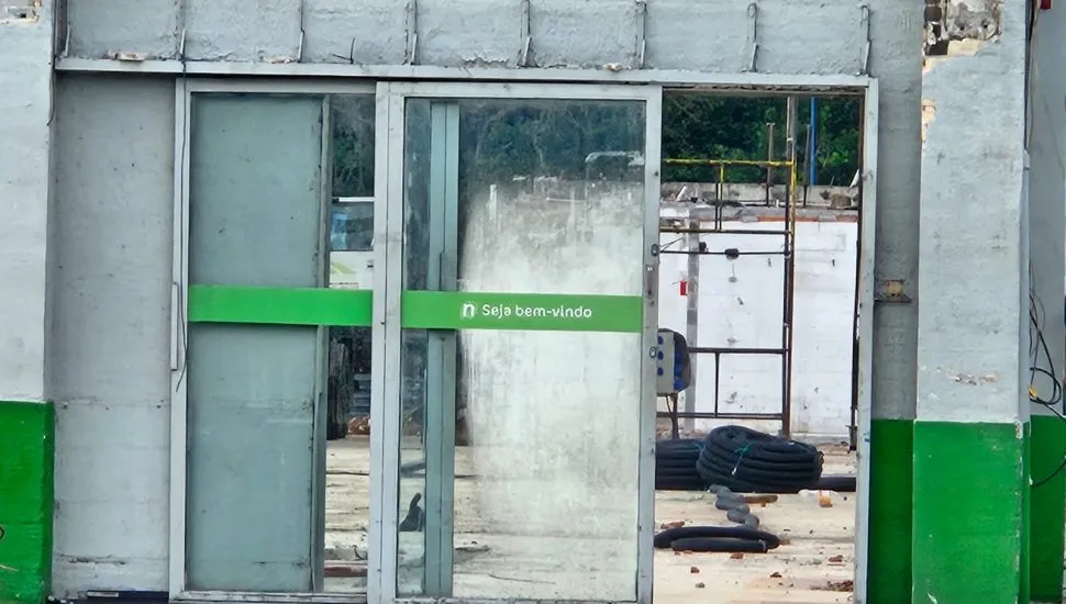 Imagem mostra porta de entrada do supermercado, com ointerior todo em obras e um adesivo na porta com a frase Seja Bem Vindo