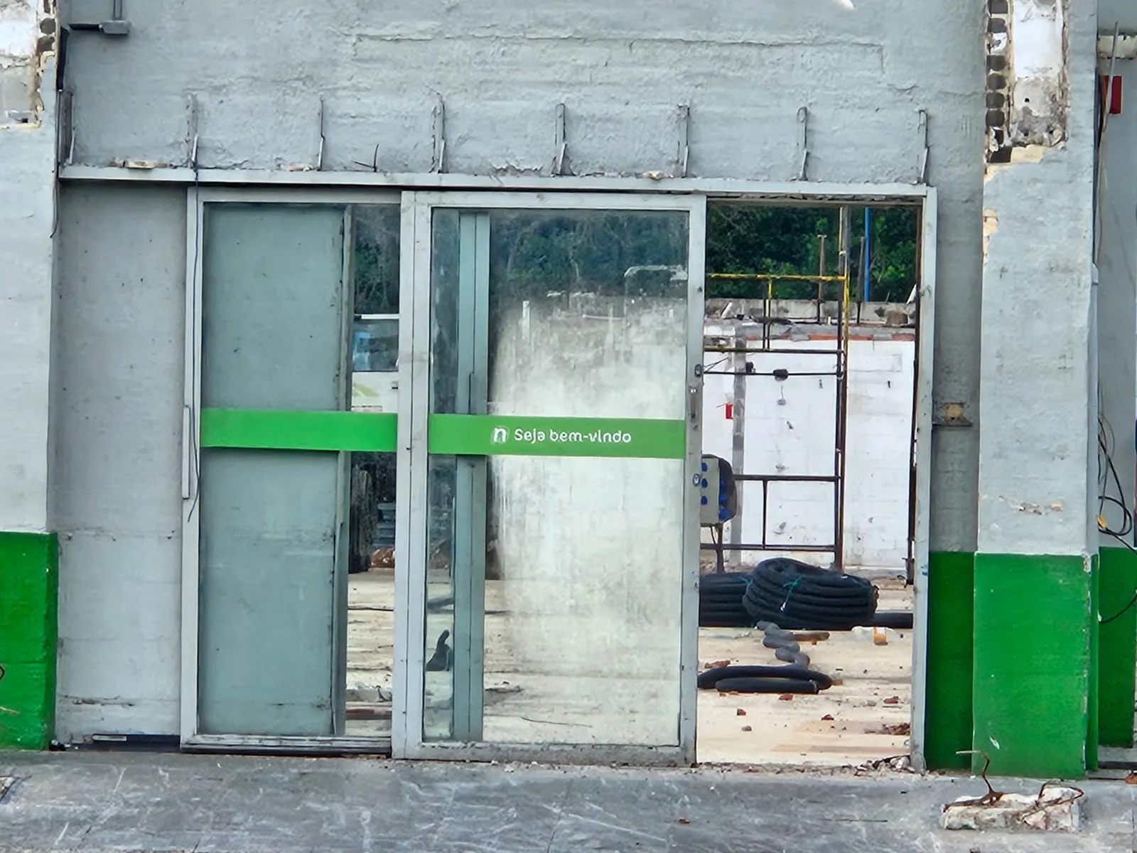 Imagem mostra porta de entrada do supermercado, com ointerior todo em obras e um adesivo na porta com a frase Seja Bem Vindo