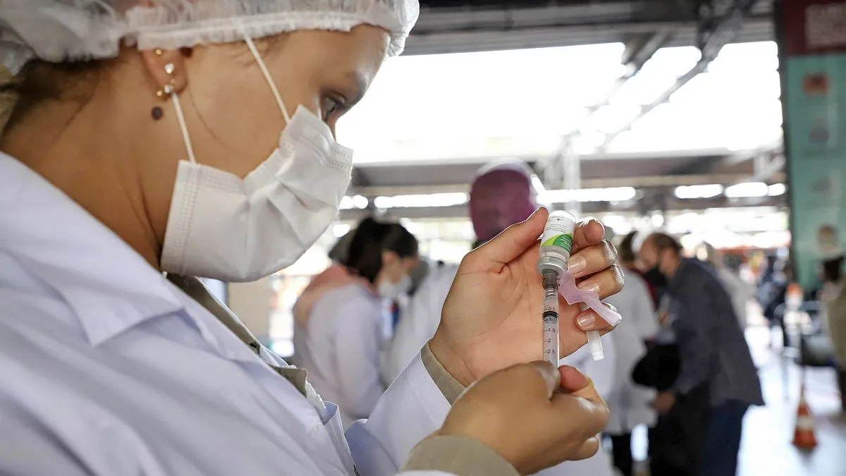 Funcionária abastece uma seringa de vacina contra a gripe, com várias pessoas ao fundo, em um terminal de ônibus