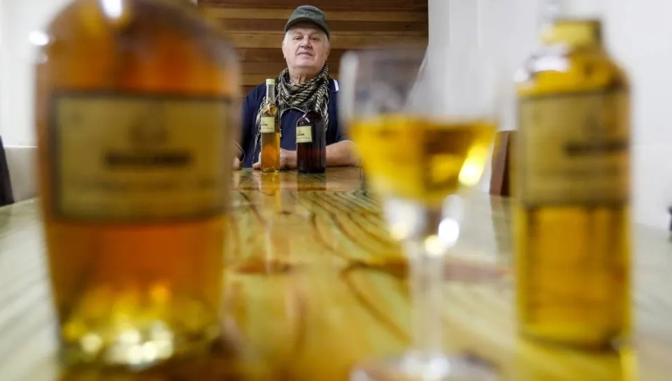 Imagem mostra uma garrafa e uma taça servida com cachaça de cataia. Ao fundo está o criador.