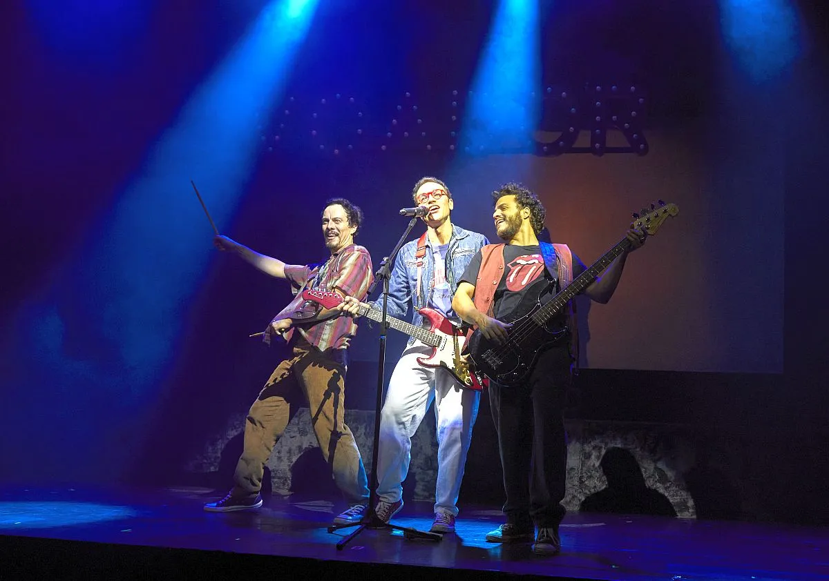 Imagem mostra os três atores que representam os paralamas no palco, dividindo um microfone