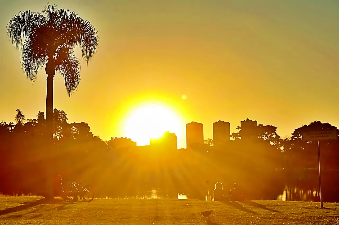 Foto mostra o por do sol, por entre prédios e pessoas correndo no Parque Barigui. Lago ao fundo