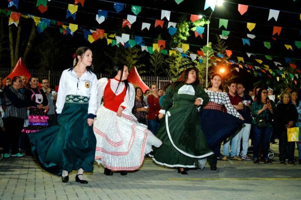 Imagem mostra uma clássica festa junina de igreja com mulheres de vestido dançando debaixo de bandeirinhas.