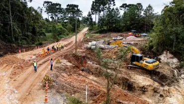 Imagem mostra estrada de chão e vários trabalhadores e máquinas fazendo o trabalho de pavimentação