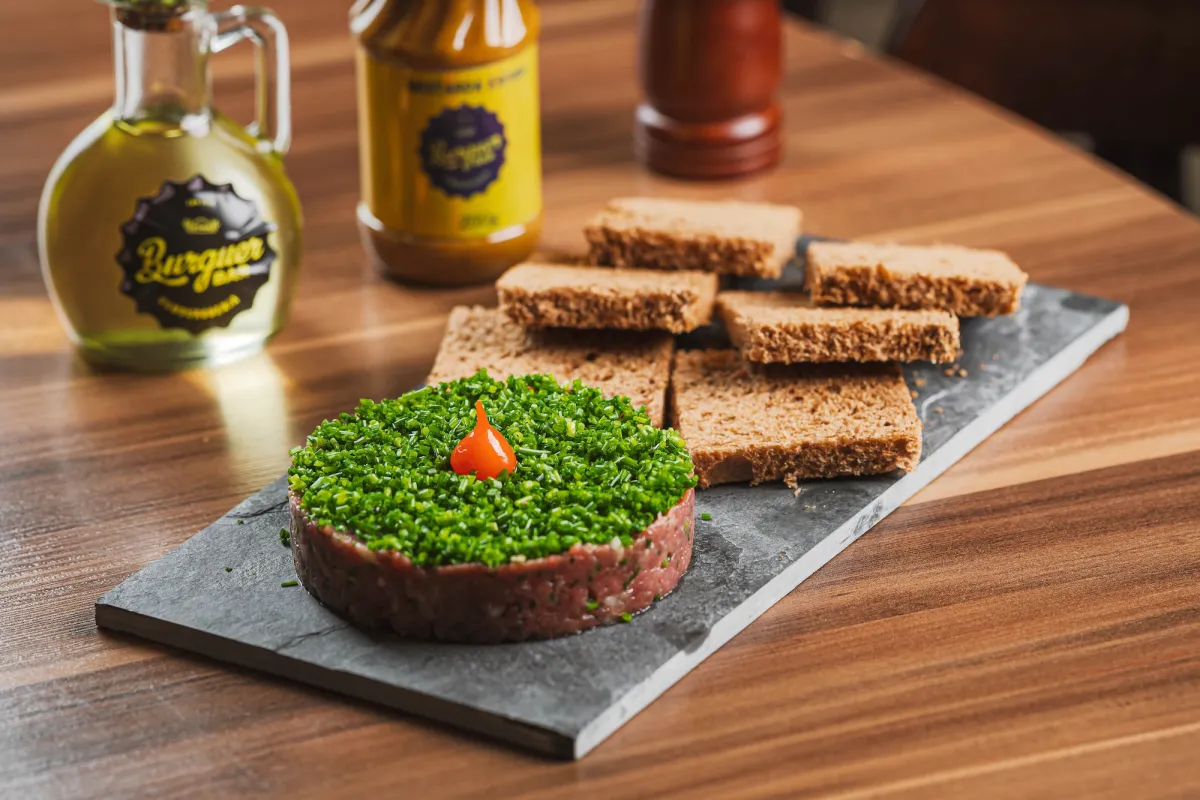 imagme msotra preparo clássico da carne de onça, com carne moída em forma de disco, fatias de broa e uma garrafa de azeite de oliva ao fundo