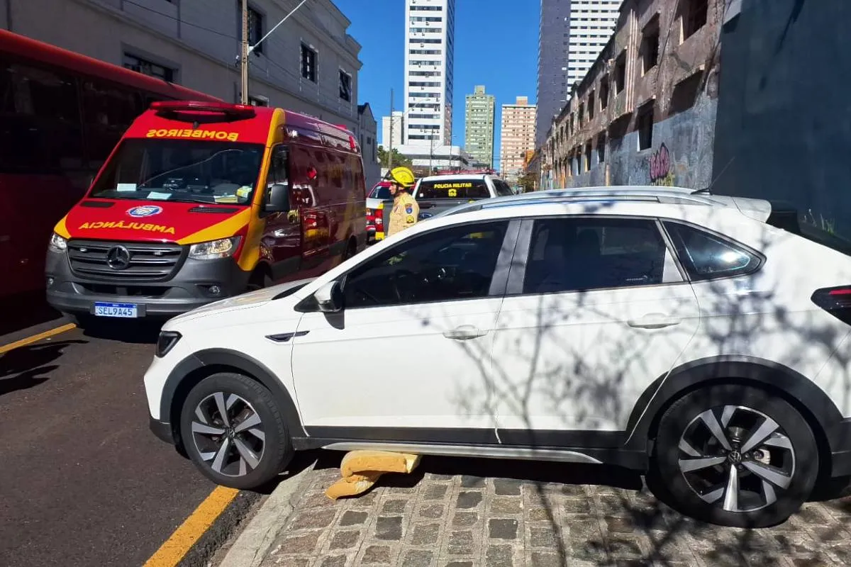 Imagem mostra um carro branco com pedaço de colhão em baixo. Há uma ambulância ao fundo