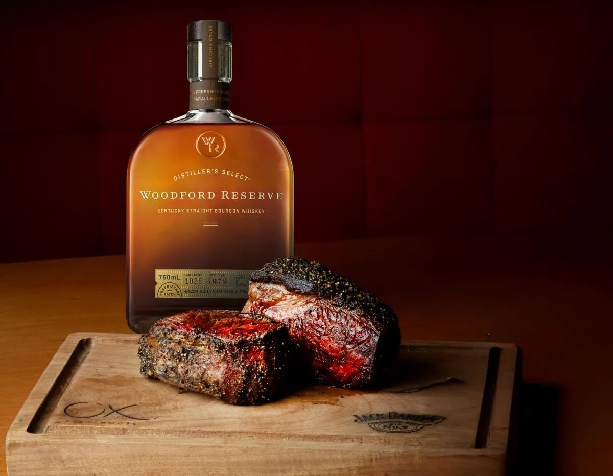 Imagem mostra um corte alto de carne bovina assada numa tábua, com garrafa de whisky ao fundo