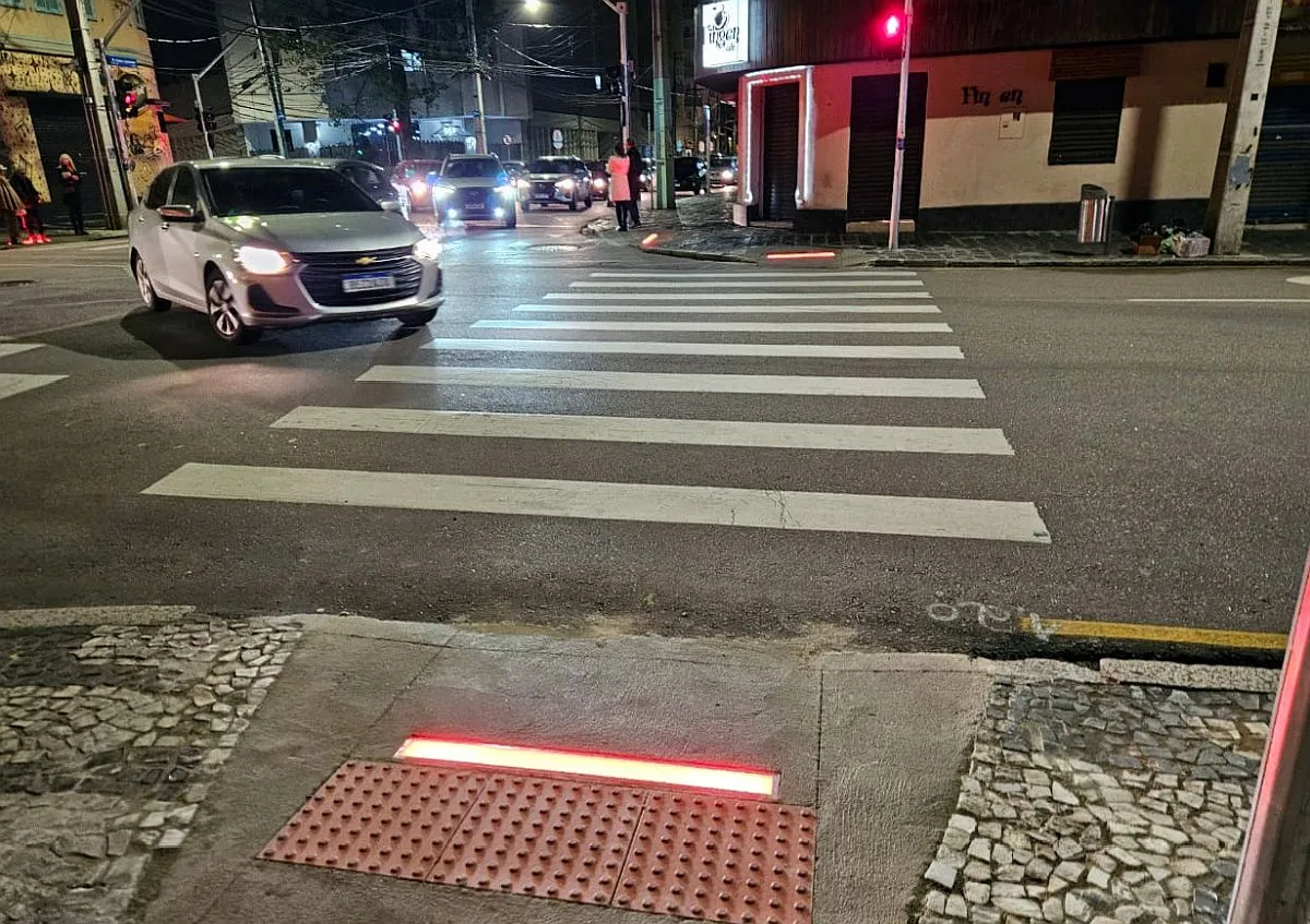 Imagem mostra cruzamento, com carros passando e um semáfogo vermelho para pedestres. A barra iluminada de vermelho está em primeiro plano