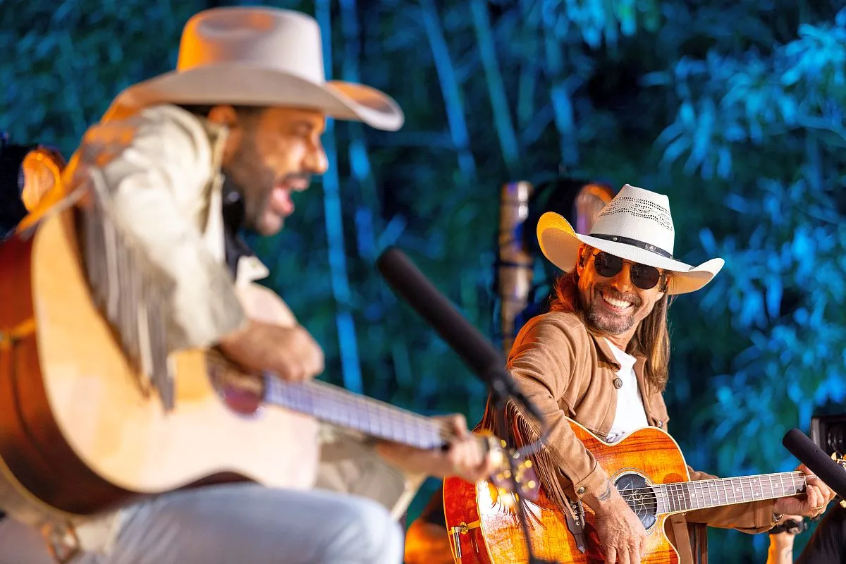 Imagem mostra irmãos Edson, em primeiro plano, mas desfocado, e Hudson ao fundo olhando para o irmão e tocando violão.