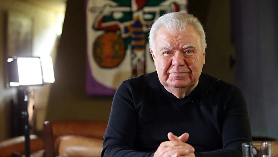 Foto mostra o ex-prefeito de Curitiba olhando para a imagem, nm escritório, com um quadro cm obra de Poty Lazarotto ao fundo