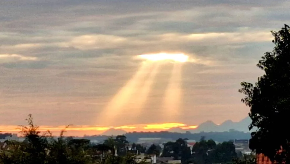 Imagem mostra raios de sol surgindo entre as nuvens com a Serra do Mar ao fundo.