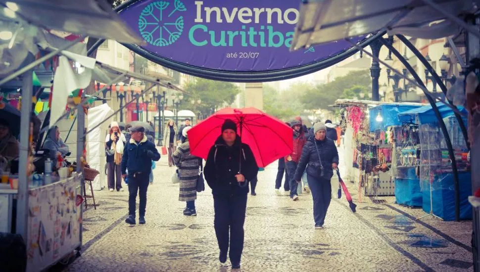 Imagem mostra uma pessoa com guarda chuva vermelho andando no Centro de Curitiba bem abaixo da placa que está escrito: Feira de inverno de Curitiba