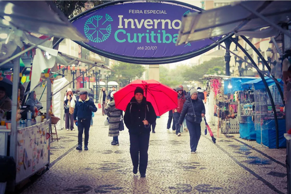 Imagem mostra uma pessoa com guarda chuva vermelho andando no Centro de Curitiba bem abaixo da placa que está escrito: Feira de inverno de Curitiba