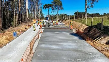 Imagem mostra pavimentação de estrada rural com concreto, cercada de árvores e cercas de fazendas da região. Pelo menos 10 trabalhadores estão no serviço