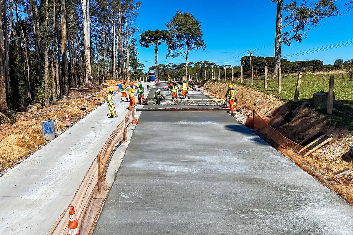 Imagem mostra pavimentação de estrada rural com concreto, cercada de árvores e cercas de fazendas da região. Pelo menos 10 trabalhadores estão no serviço