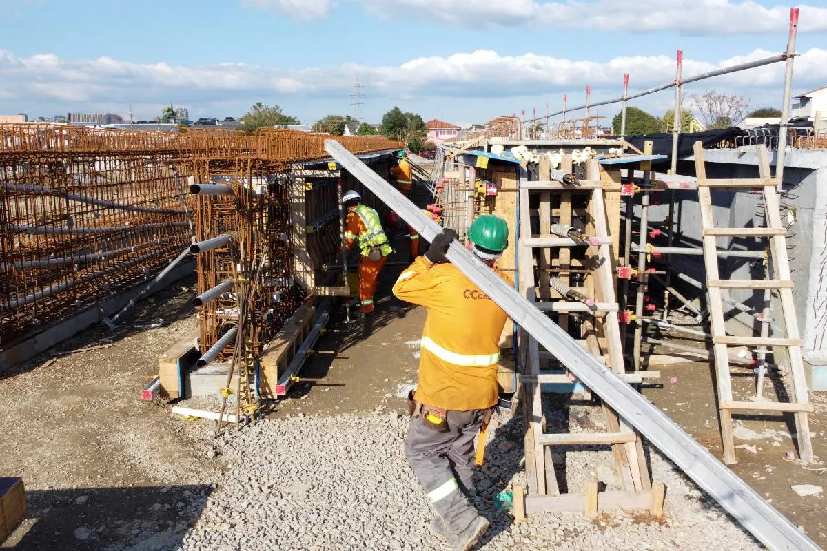 Imagem mostra um trabalhador carregando uma viga em uma obra.