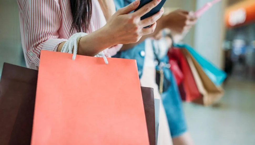 Imagem mostra mulher segurando diversas sacolas e usando o celular