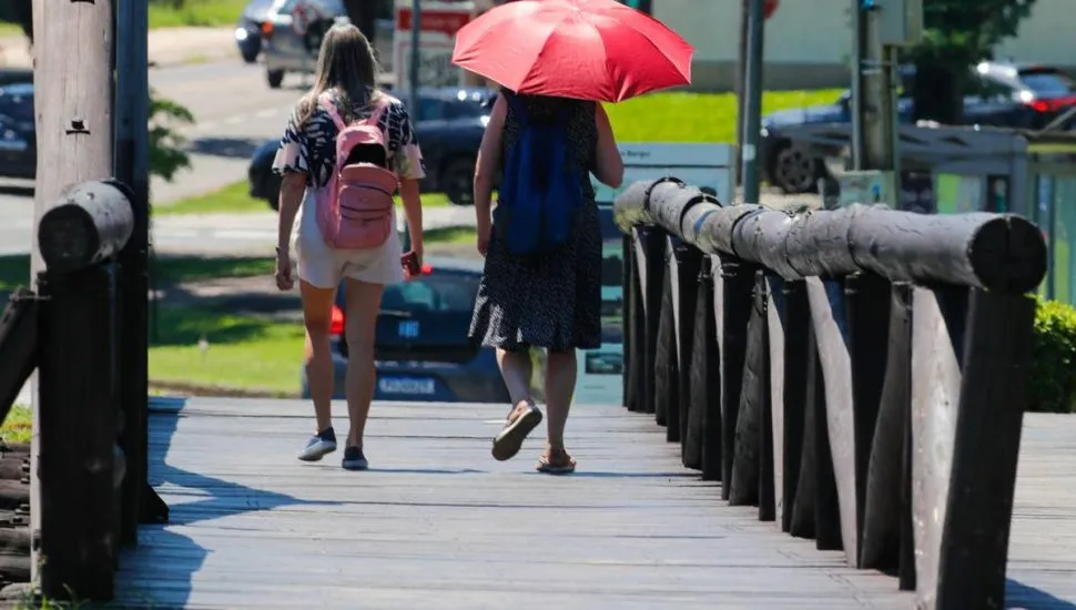 Imagem mostra pessoas com calor andando em um parque. Uma pessoa se protege do sol com um guarda chuva.